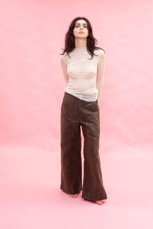 A woman stands against a solid pink studio backdrop wearing a sheer cream long-sleeve top made from stretch knitted silk tulle,and dark brown wide-leg corduroy pants.
