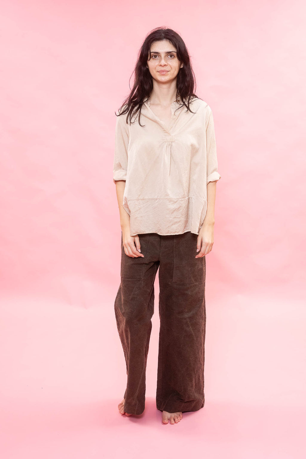 Woman wearing a beige blouse and brown pants against a pink background