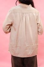 A person viewed from the back wearing a light beige long-sleeve blouse with a gathered back neckline and dark brown pants, set against a solid pink background.