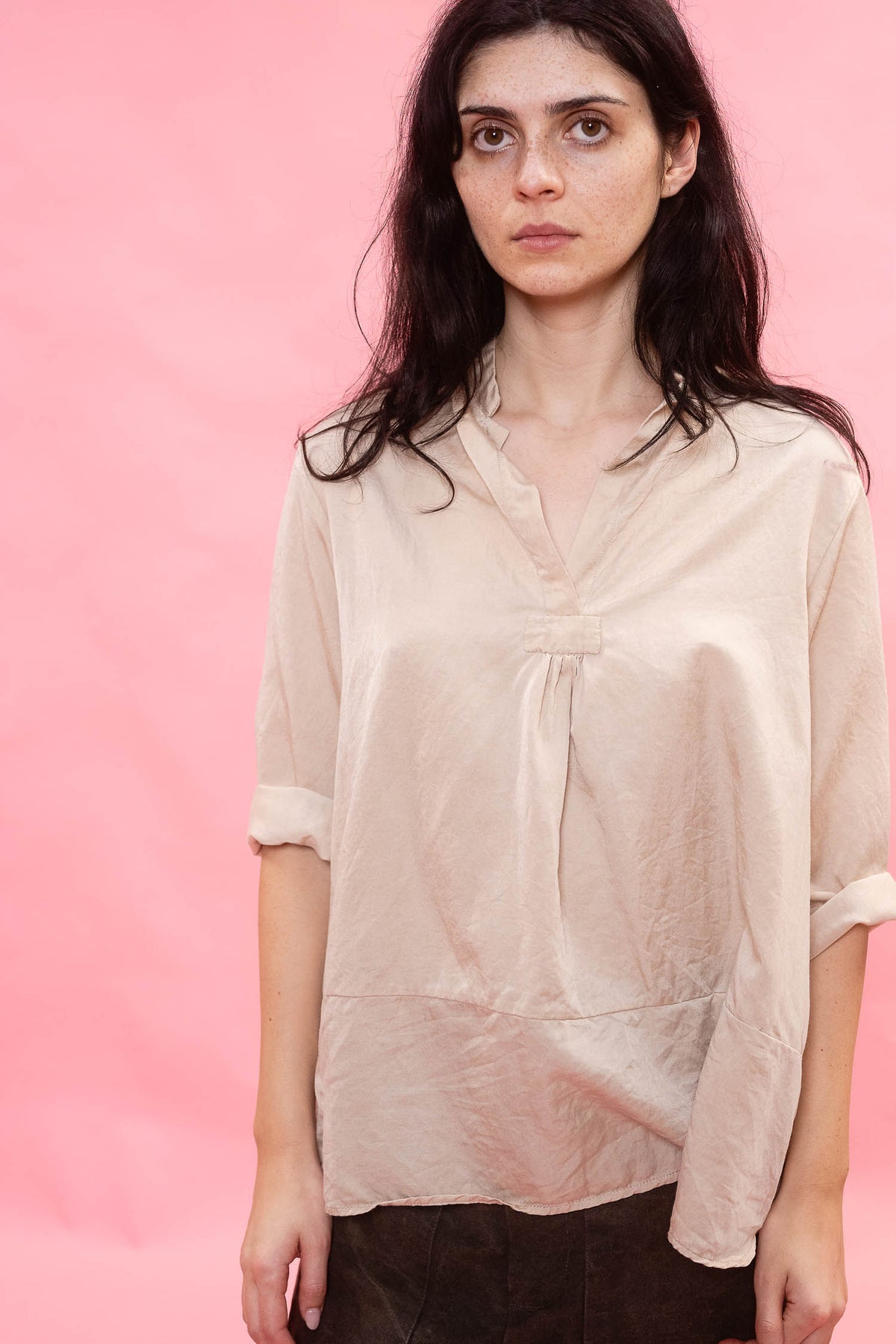 A woman modeling a loose, light beige linen blouse with a gathered front and rolled sleeves, set against a solid pink background.
