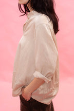 A woman shown from the side wearing a loose, light beige linen button-up shirt with rolled-up sleeves, paired with dark brown pants, set against a solid pink background.