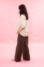 A woman stands barefoot in profile against a solid pink studio backdrop, wearing a loose off-white blouse and dark brown wide-leg corduroy pants.