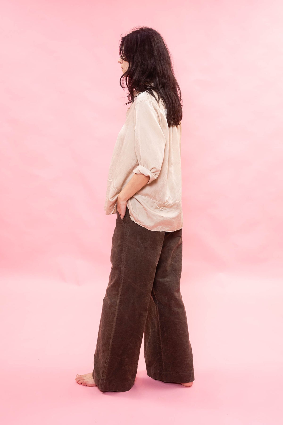 A woman stands barefoot in profile against a solid pink studio backdrop, wearing a loose off-white blouse and dark brown wide-leg corduroy pants.