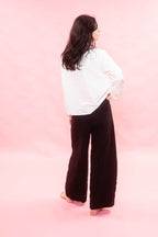 A woman is shown from behind wearing a white long-sleeve blouse and dark brown wide-leg pleated trousers, standing against a solid pink studio background.