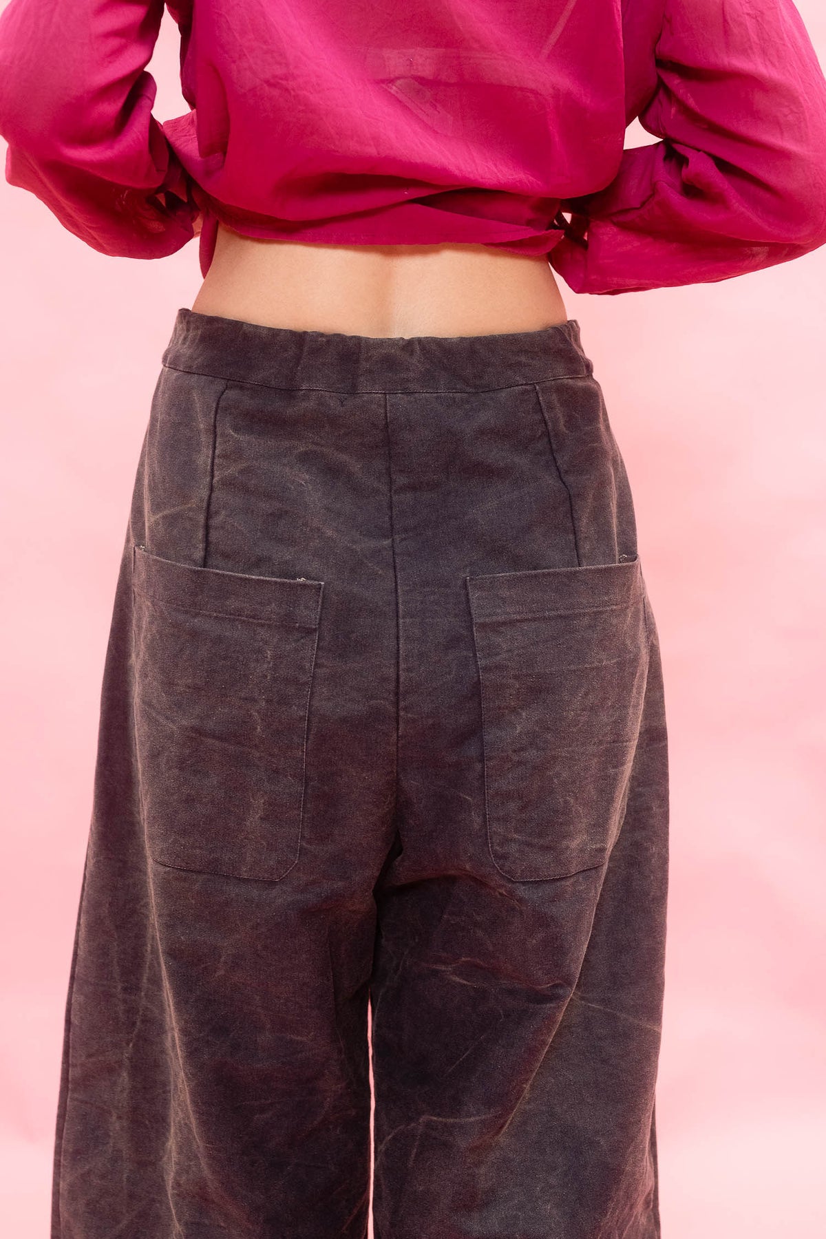 A person shown from the back wearing dark charcoal wide-leg trousers with large patch pockets and a bright pink cropped long-sleeve top, against a solid pink background.