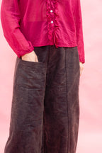 A person wearing a bright pink ruffled blouse and dark brown wide-leg trousers, shown from the chest down against a soft pink studio background.