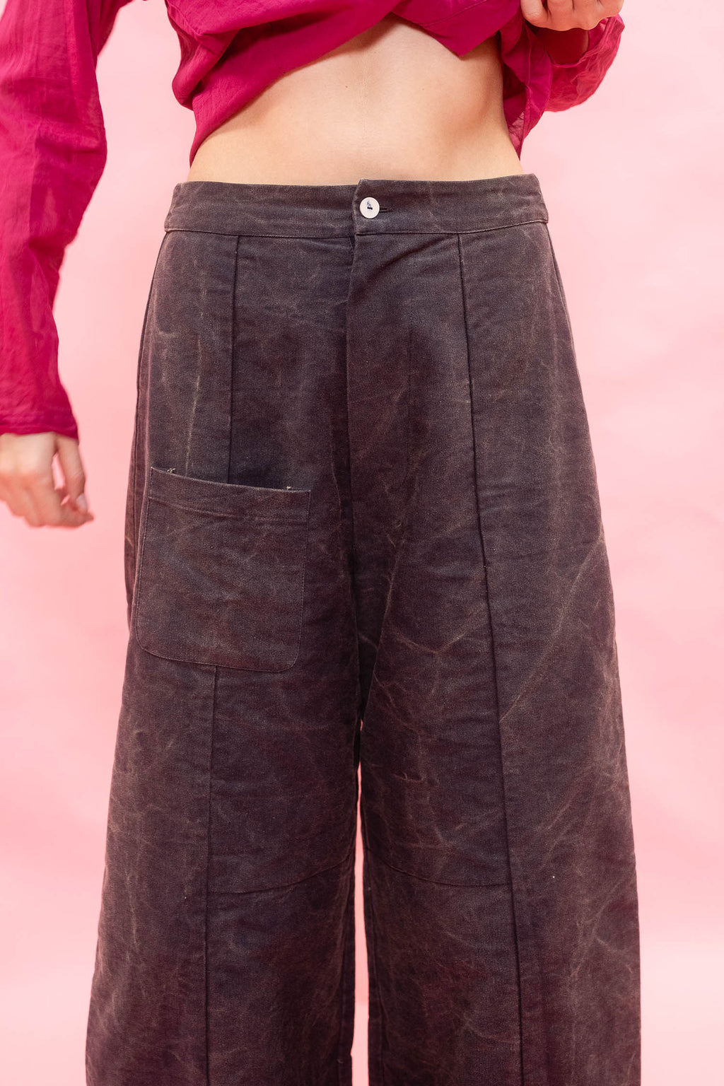 A person wearing dark brown wide-leg corduroy pants with a button waistband and front patch pocket, paired with a magenta cropped top, against a solid pink background.