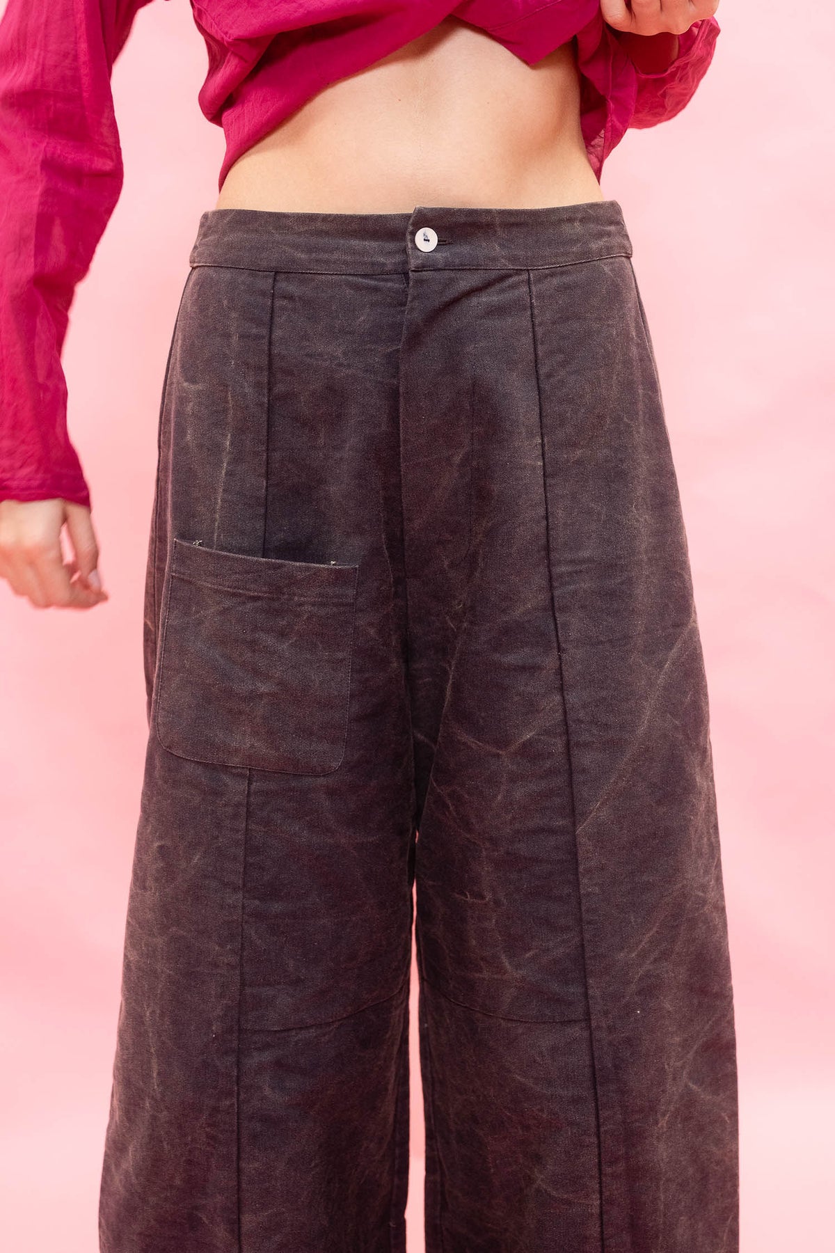 A person wearing dark brown wide-leg corduroy pants with a button waistband and front patch pocket, paired with a magenta cropped top, against a solid pink background.