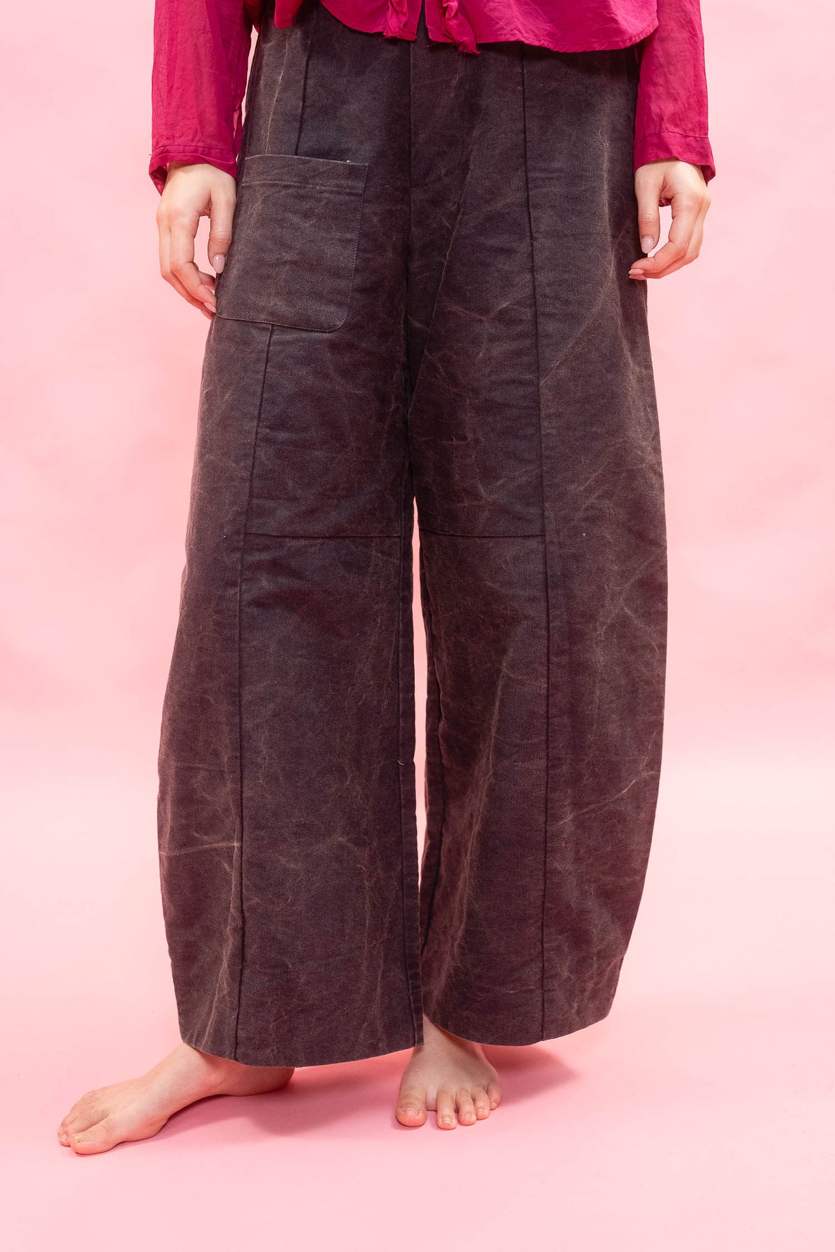 A person wearing very wide-leg dark brown pants with a textured, waxed finish, paired with a bright pink top, standing barefoot against a solid pink background.