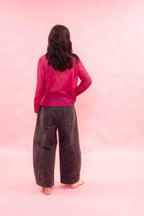 A woman viewed from behind wearing a bright pink sheer long-sleeve top and dark charcoal gray wide-leg pants, standing against a solid pink backdrop.