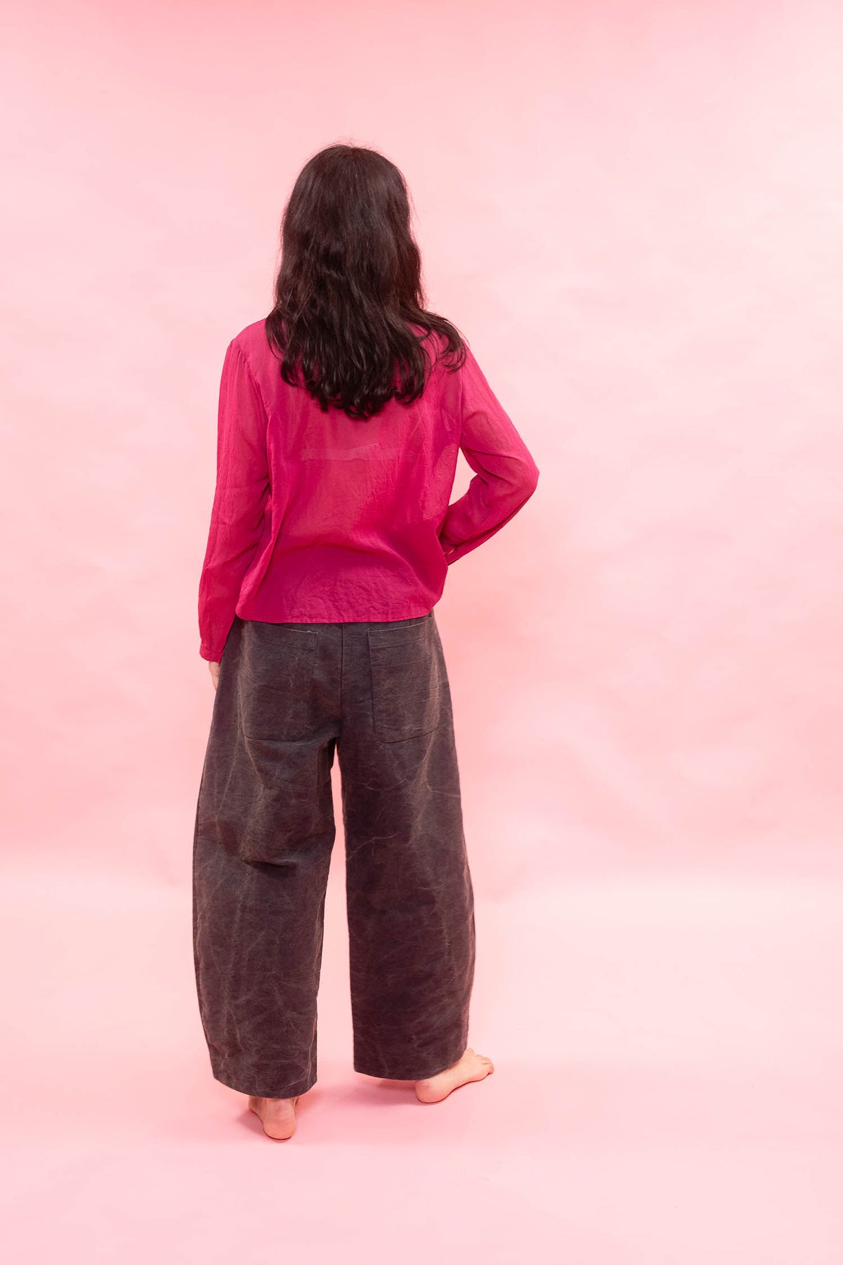 A woman viewed from behind wearing a bright pink sheer long-sleeve top and dark charcoal gray wide-leg pants, standing against a solid pink backdrop.