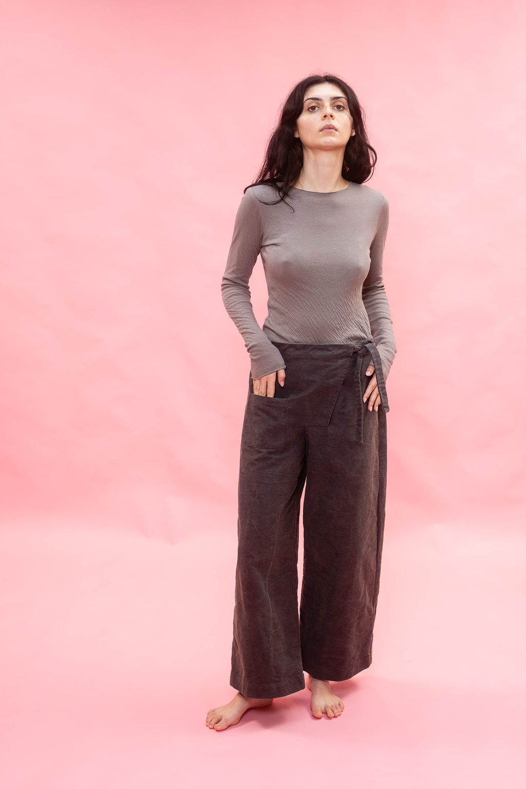 A woman stands barefoot against a solid pink backdrop, wearing a fitted long-sleeve gray top and dark navy wide-leg pants.