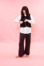 A woman stands barefoot against a solid pink studio background, wearing a white long-sleeve button-up shirt, dark navy wide-leg pants, and a black fuzzy sleeveless vest with a dark tie belt.