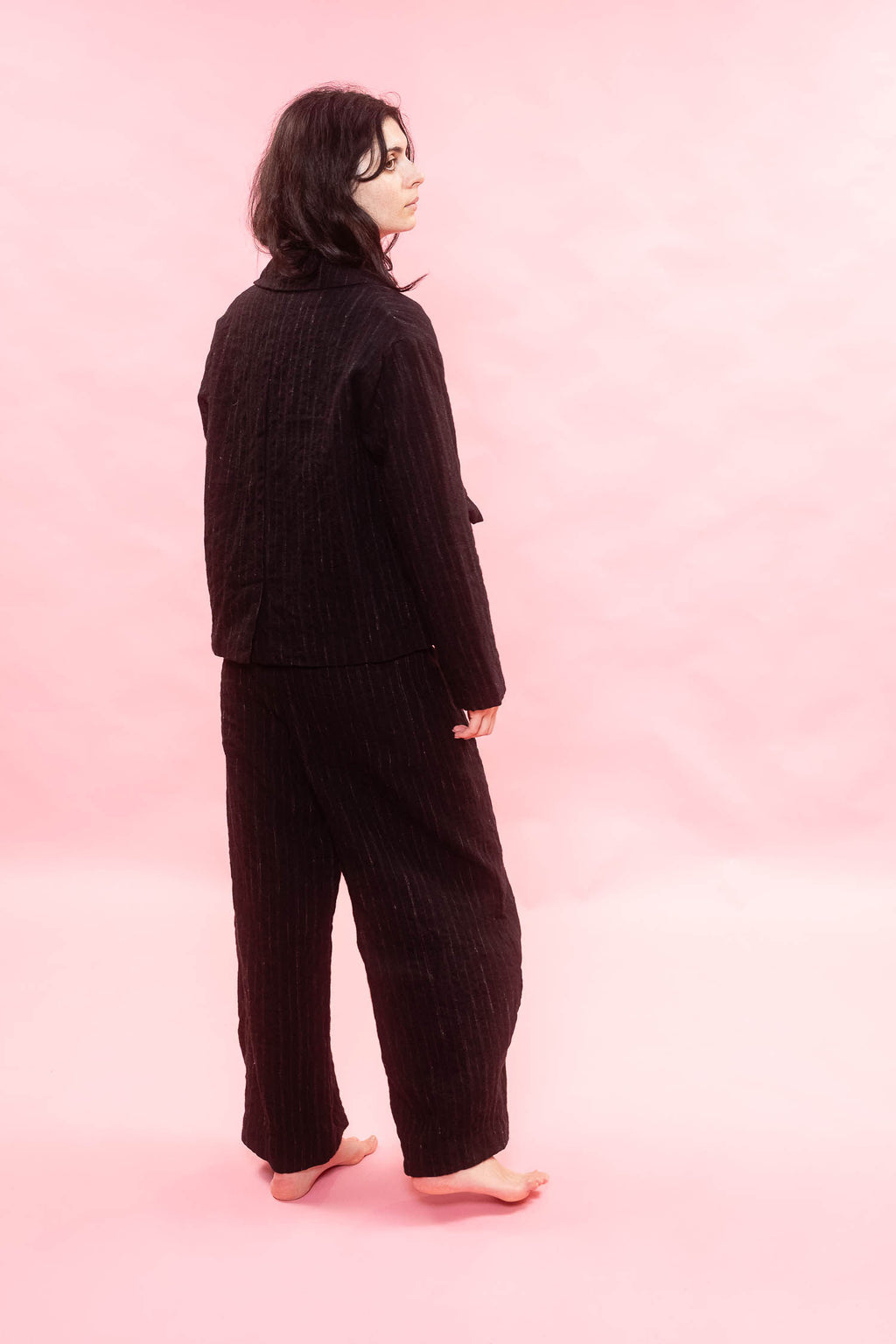 A woman standing barefoot in profile against a soft pink studio background, wearing a black pinstripe matching set consisting of a button front jacket and wide-leg pants.