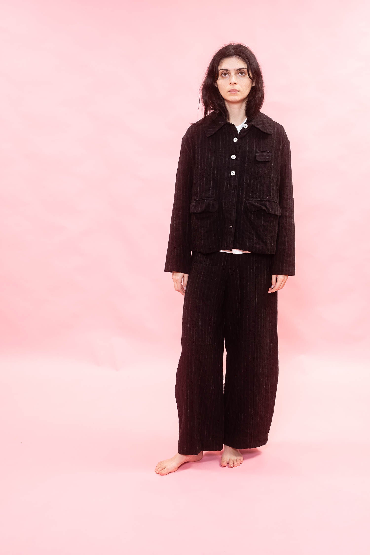 A model stands barefoot against a solid pink studio background, wearing a matching wool pinstripe two-piece set consisting of a cropped button-up jacket and wide-leg trousers named tailored line pants