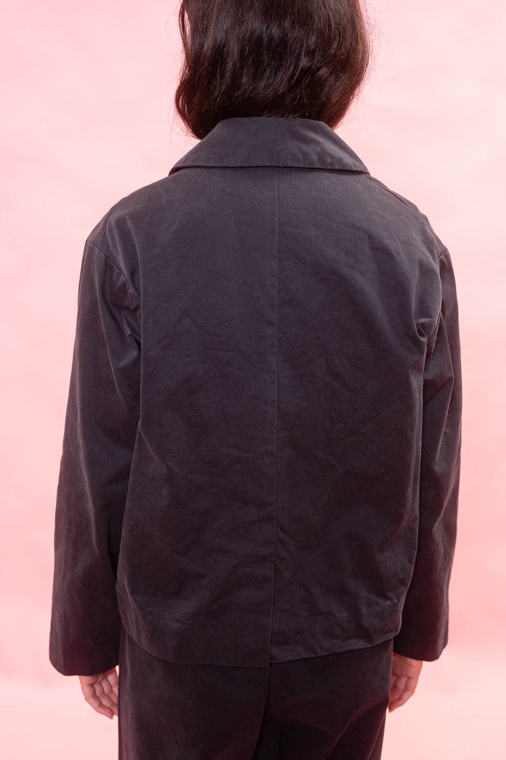 A person viewed from the back wearing a dark navy jacket and matching dark pants against a solid pink background.