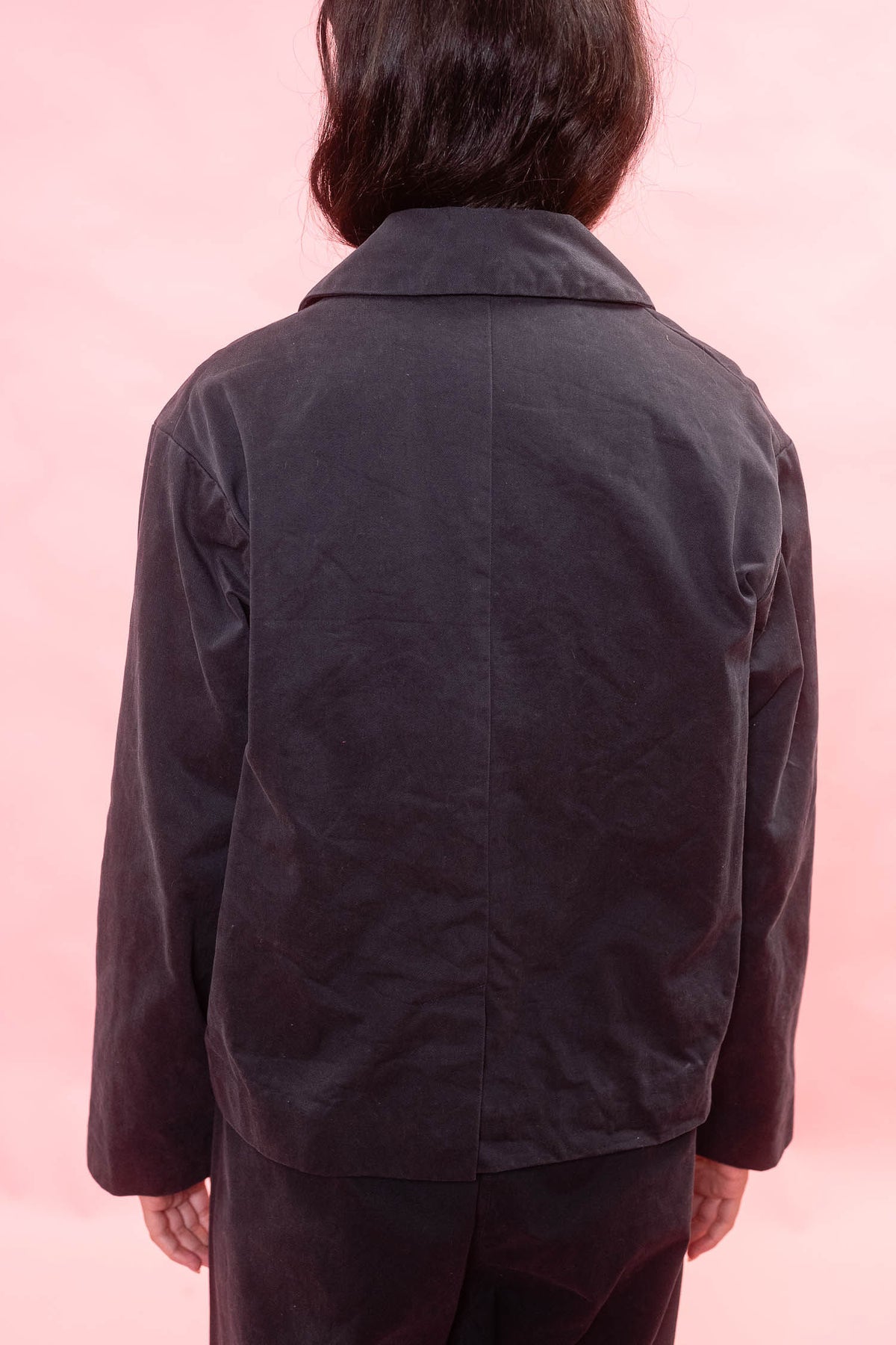 A person viewed from the back wearing a dark navy jacket and matching dark pants against a solid pink background.