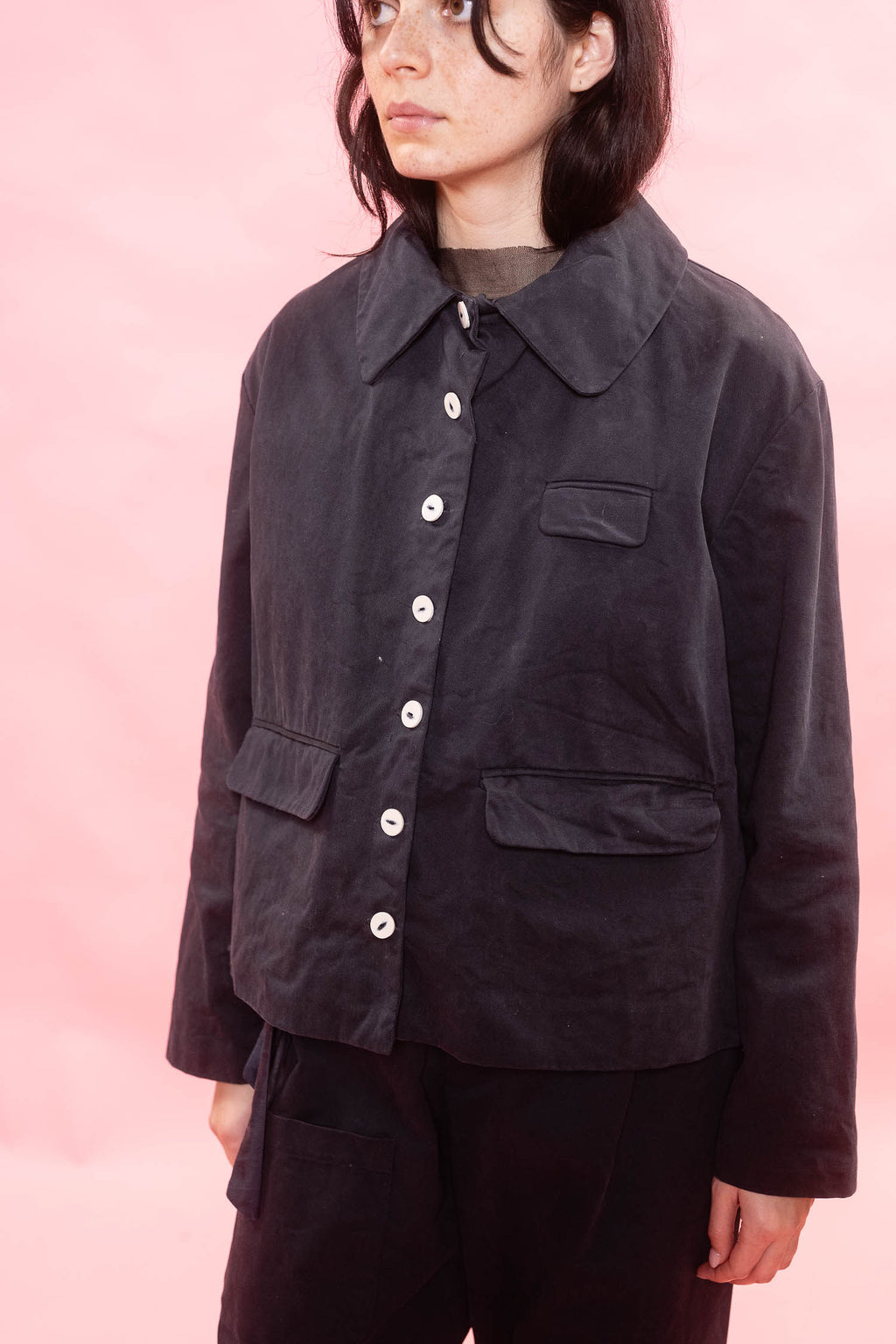 A model wearing a dark navy corduroy jacket with white buttons, paired with navy pants, set against a solid pink background.