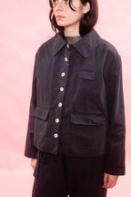 A model wearing a dark navy corduroy jacket with white buttons, paired with navy pants, set against a solid pink background.
