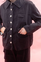 A person wearing a dark nav cotton button-up shirt jacket with a collar, chest pocket, and side pockets, paired with dark pants, set against a solid pink background.