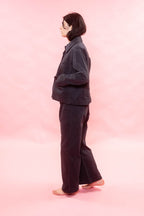 A person standing in profile wearing a dark navy jacket and matching wide-leg pants against a solid pink studio background.