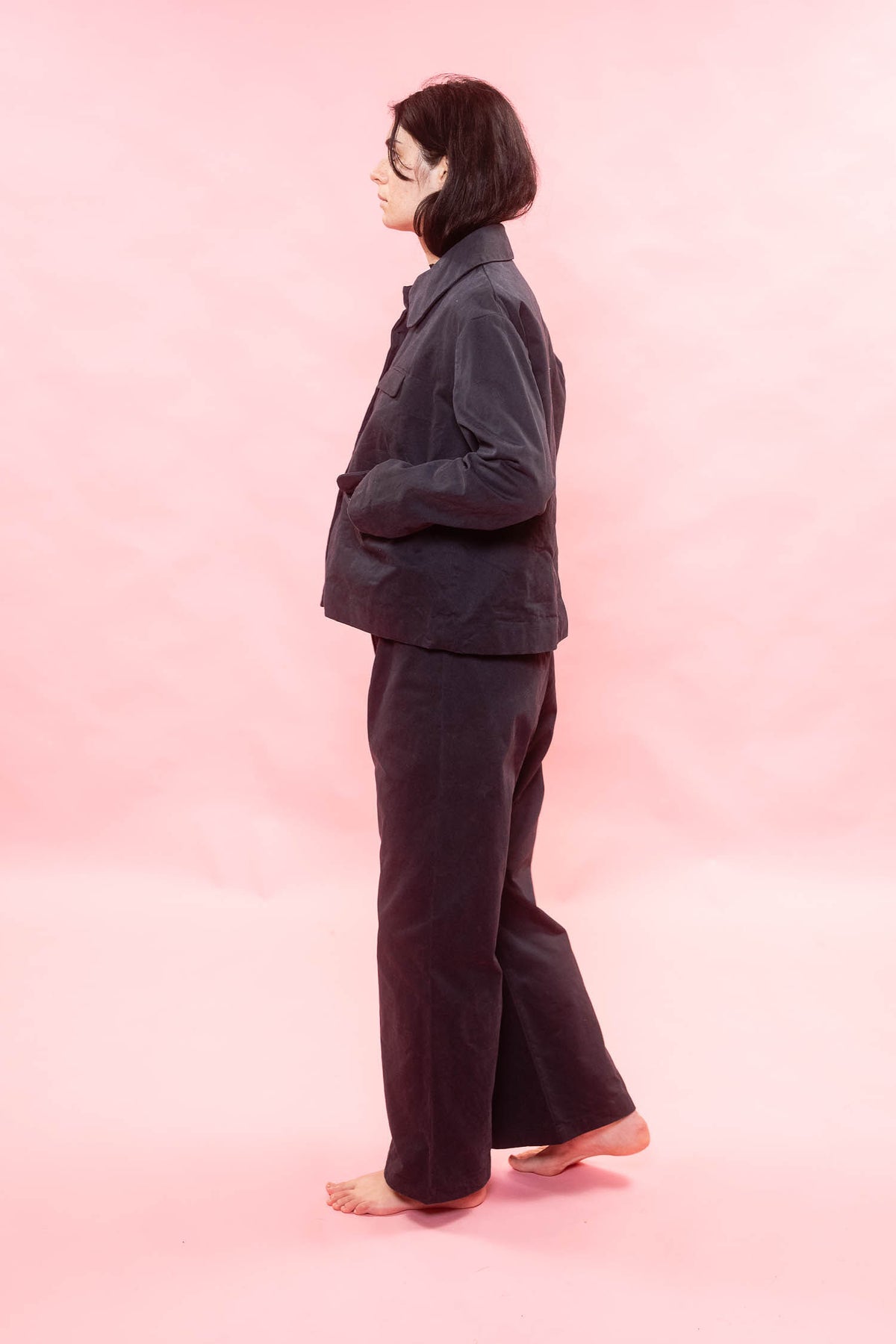 A person standing in profile wearing a dark navy jacket and matching wide-leg pants against a solid pink studio background.