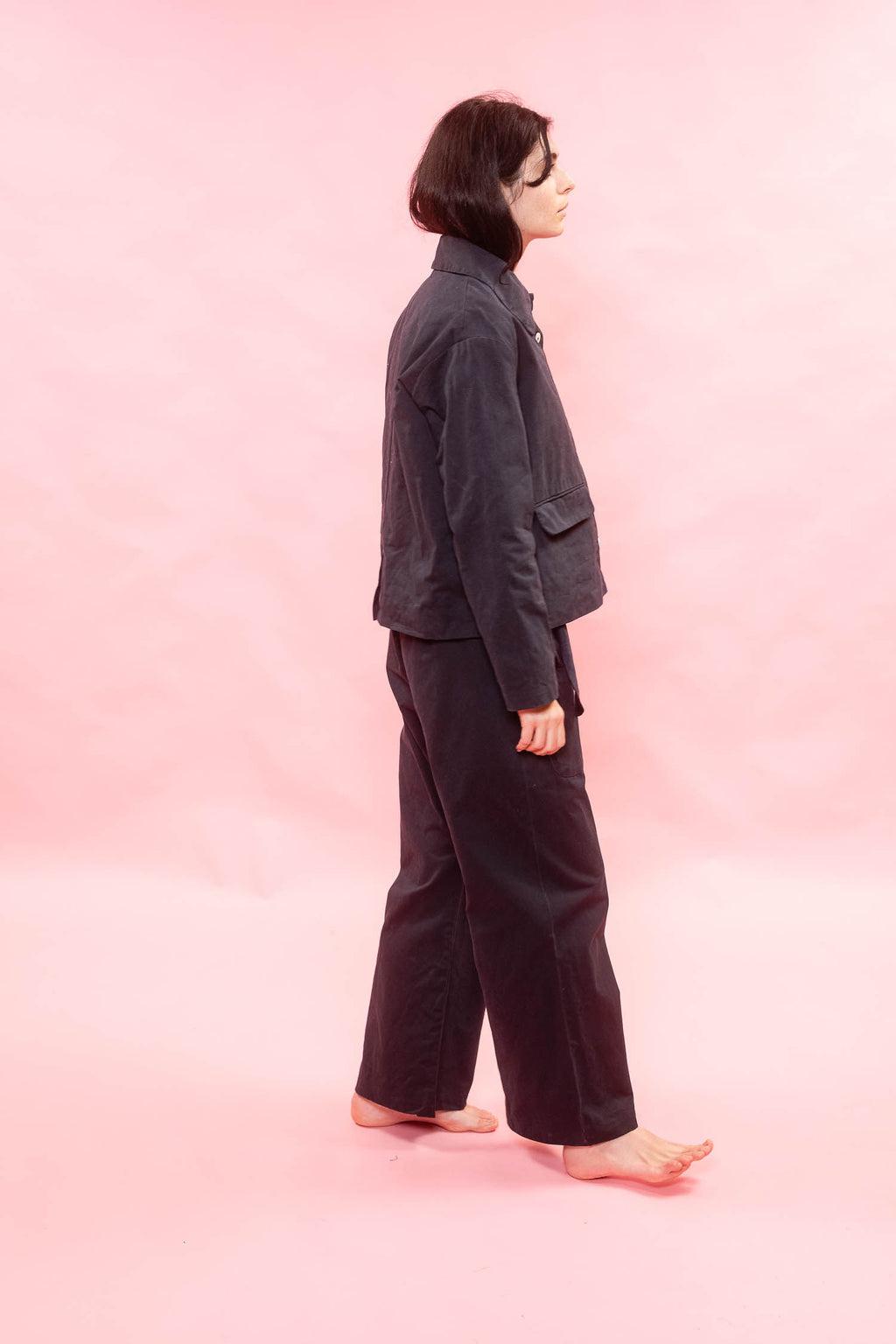 A person standing in profile against a solid pink studio background, wearing a dark navy jacket and matching dark wide-leg pants.