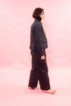A person standing in profile against a solid pink studio background, wearing a dark navy jacket and matching dark wide-leg pants.