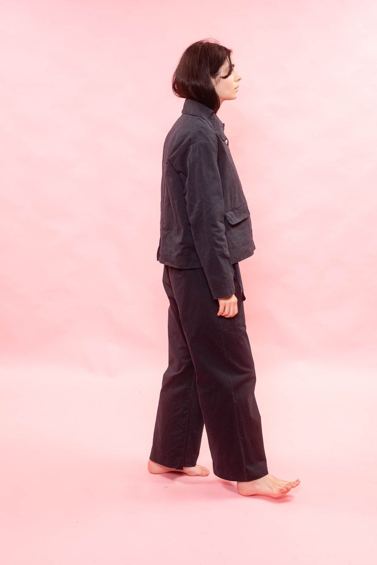 A person standing in profile against a solid pink studio background, wearing a dark navy jacket and matching dark wide-leg pants.