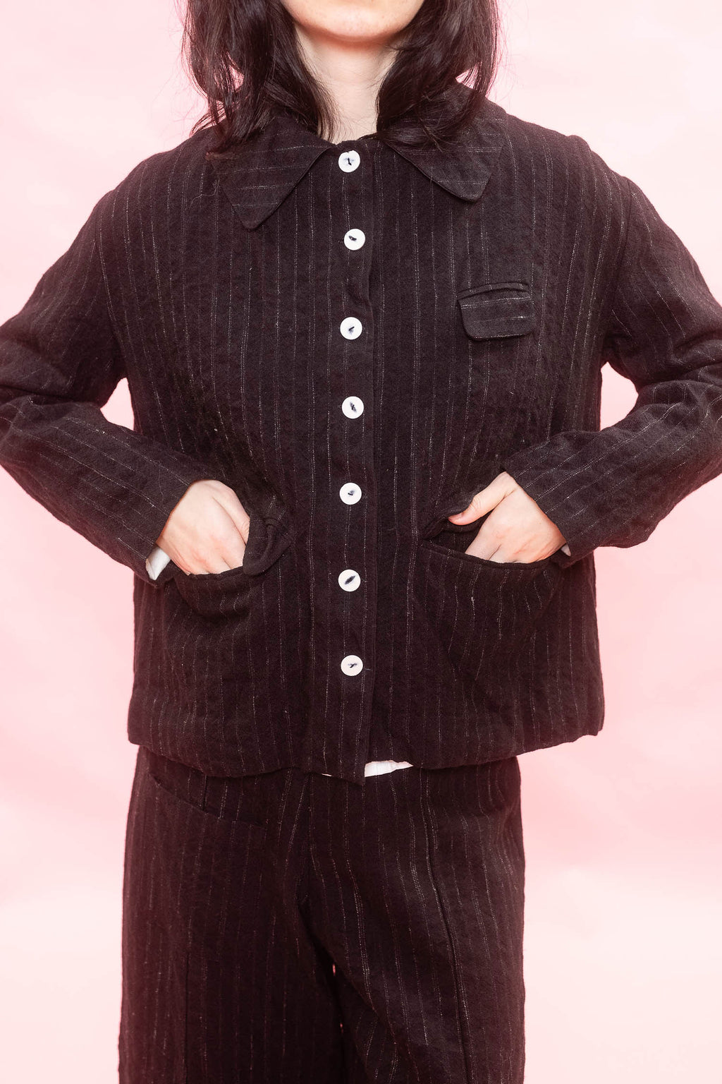 A woman wearing a black and chalk pinstripe button-front jacket with a collar and matching pinstripe tailored wide-leg pants, posed against a soft pink studio background.