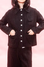 A woman wearing a black and chalk pinstripe button-front jacket with a collar and matching pinstripe tailored wide-leg pants, posed against a soft pink studio background.
