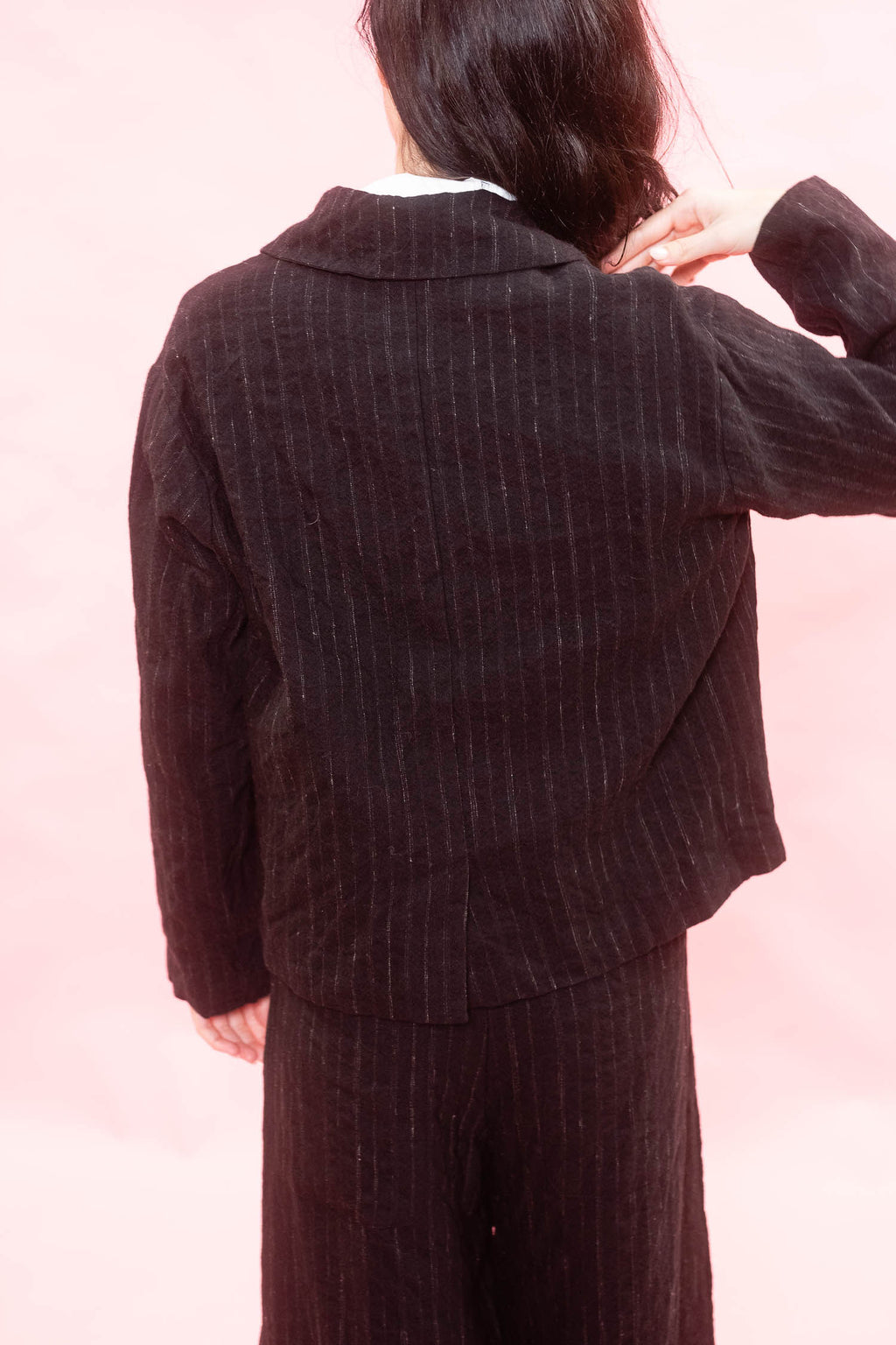 A woman shown from the back wearing a black pinstriped blazer and matching trousers against a soft pink background.