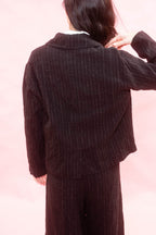A woman shown from the back wearing a black pinstriped blazer and matching trousers against a soft pink background.