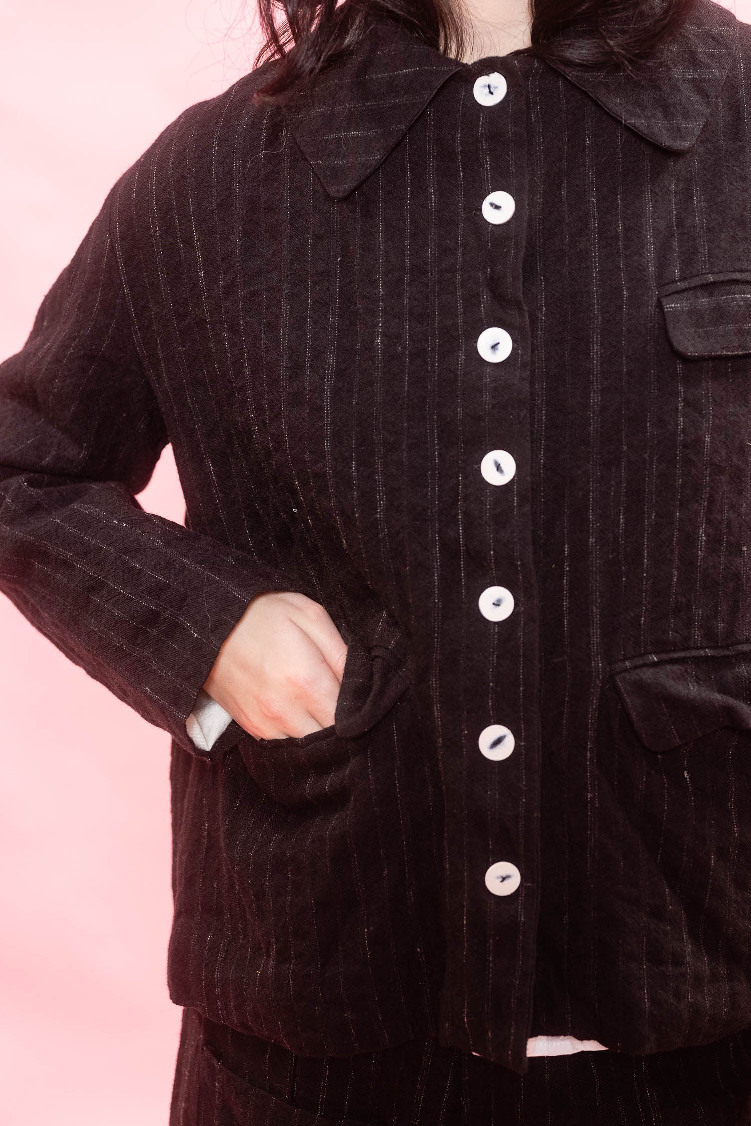 A close-up of a woman wearing a black pinstripe wool and linen jacket with white buttons, set against a solid pink background.