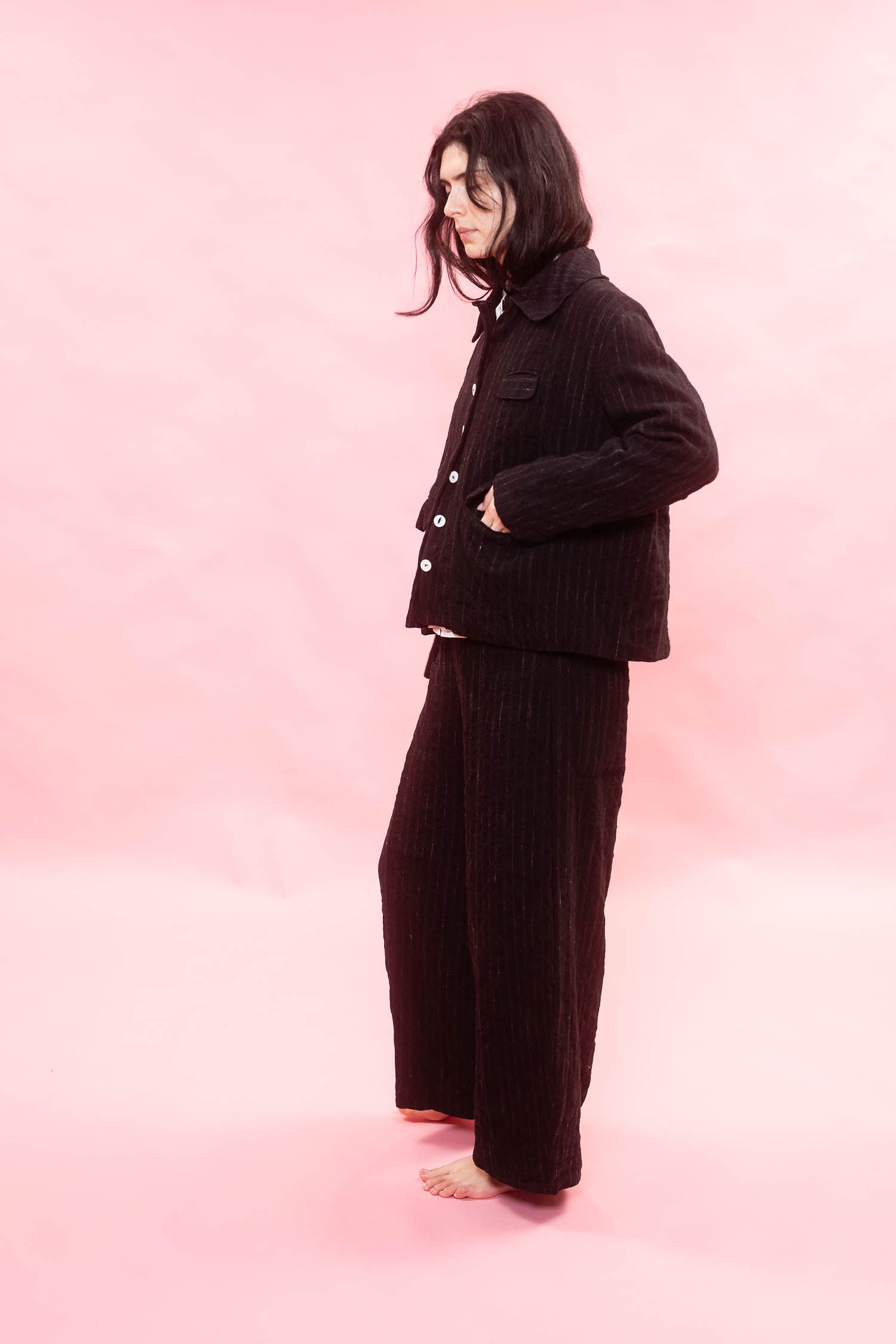 A model stands barefoot against a solid pink studio backdrop, wearing a matching black and chalk pinstripe button up jacket and wide-leg trousers.