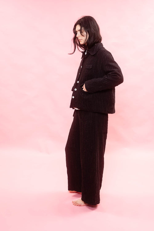 A model stands barefoot against a solid pink studio backdrop, wearing a matching black and chalk pinstripe button up jacket and wide-leg trousers.