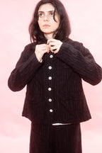 A model wearing a black and white broken pinstripe wool and linen jacket with white buttons, posed against a solid pink background.