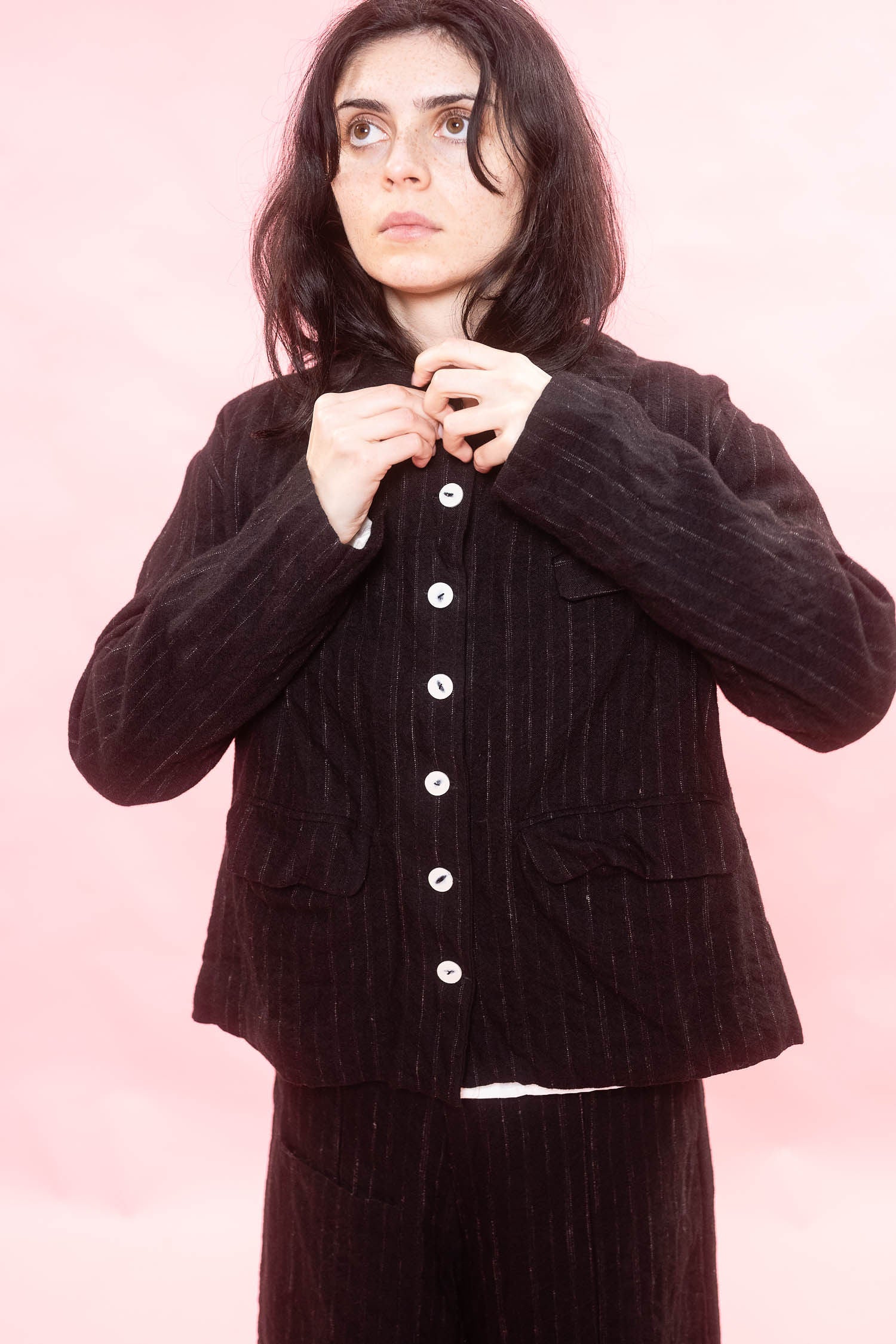 A model wearing a black and white broken pinstripe wool and linen jacket with white buttons, posed against a solid pink background.