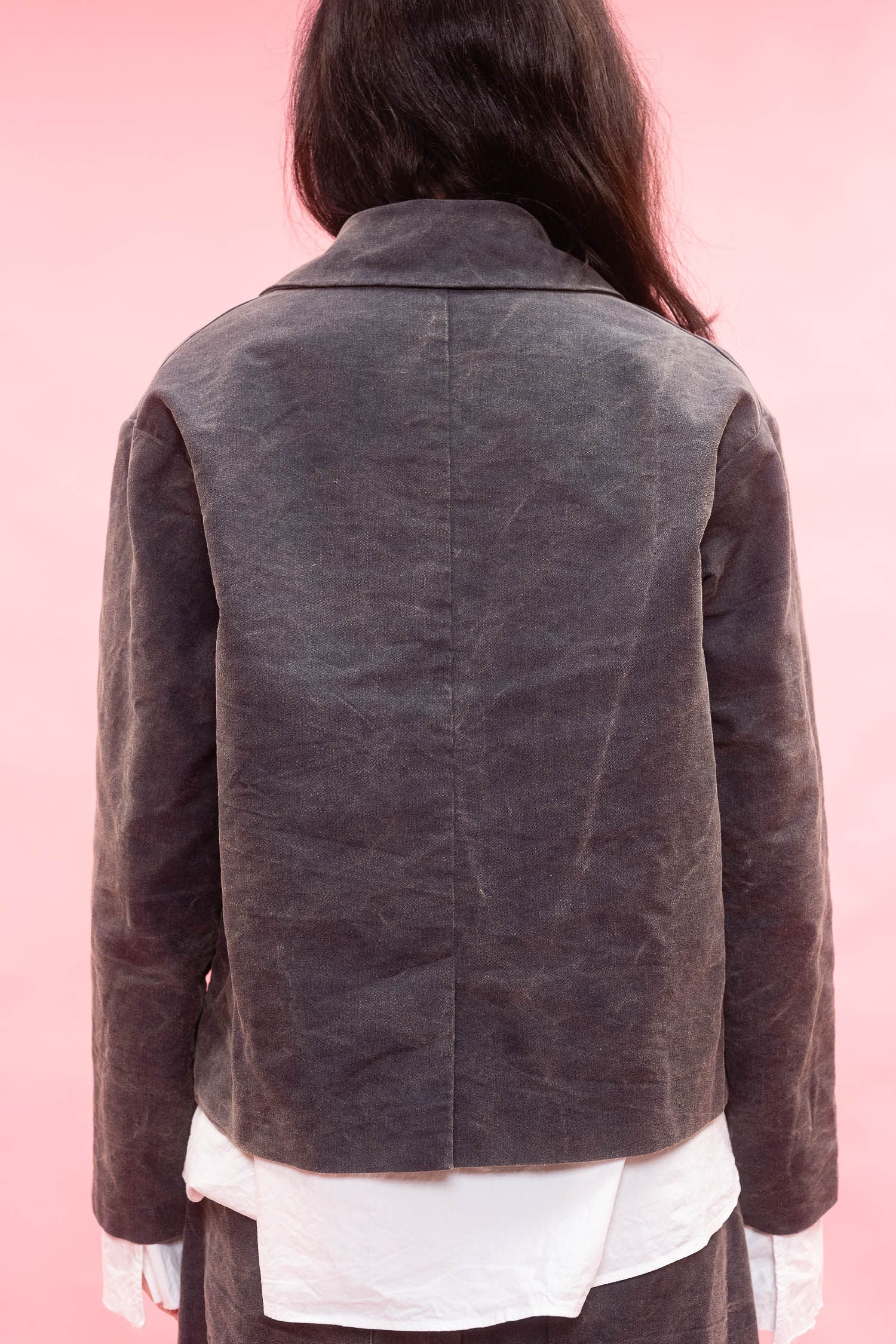 A person shown from the back wearing a grey textured jacket over a white shirt, set against a solid pink background.