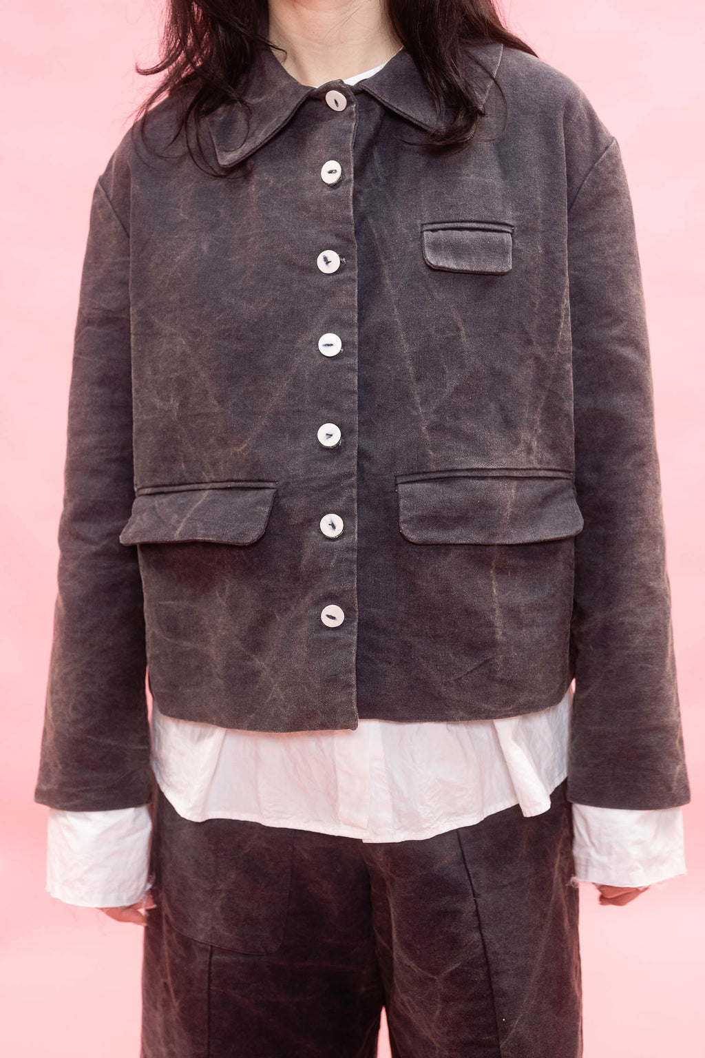 A person wearing a dark  washed cotton jacket with white shirt and dark brown pants, set against a solid pink background.