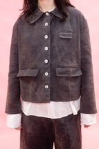 A person wearing a dark  washed cotton jacket with white shirt and dark brown pants, set against a solid pink background.