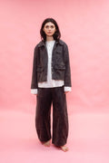 A model stands barefoot against a solid pink studio backdrop, wearing a dark brown corduroy jacket over a white shirt and matching dark brown corduroy wide-leg trousers.