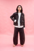 A woman stands barefoot against a solid pink backdrop, wearing a dark washed cotton jacket and matching wide-leg trousers over a white button-up shirt.