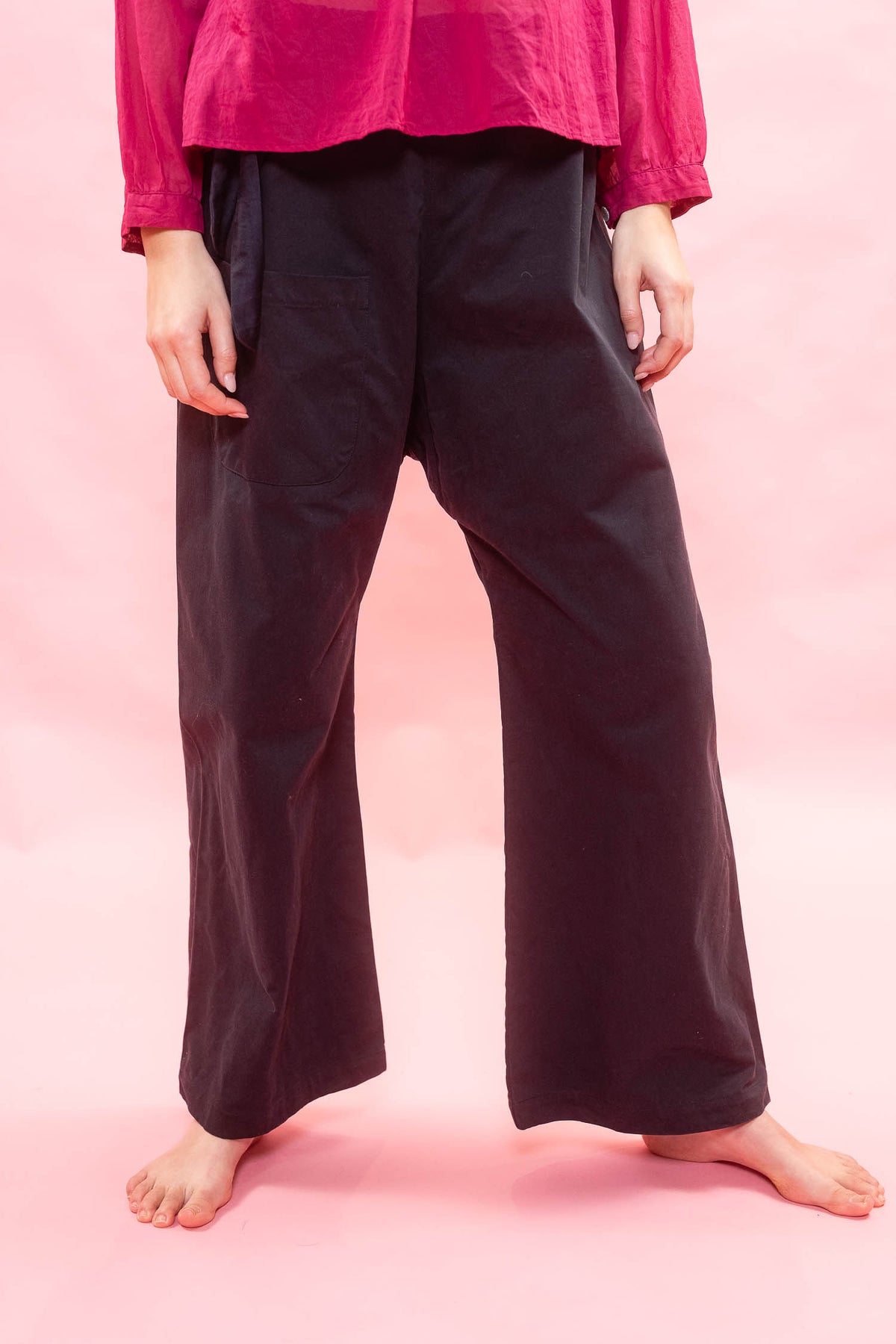 A person wearing loose, wide-leg navy pants and a bright pink long-sleeve top, standing barefoot against a solid pink backdrop.