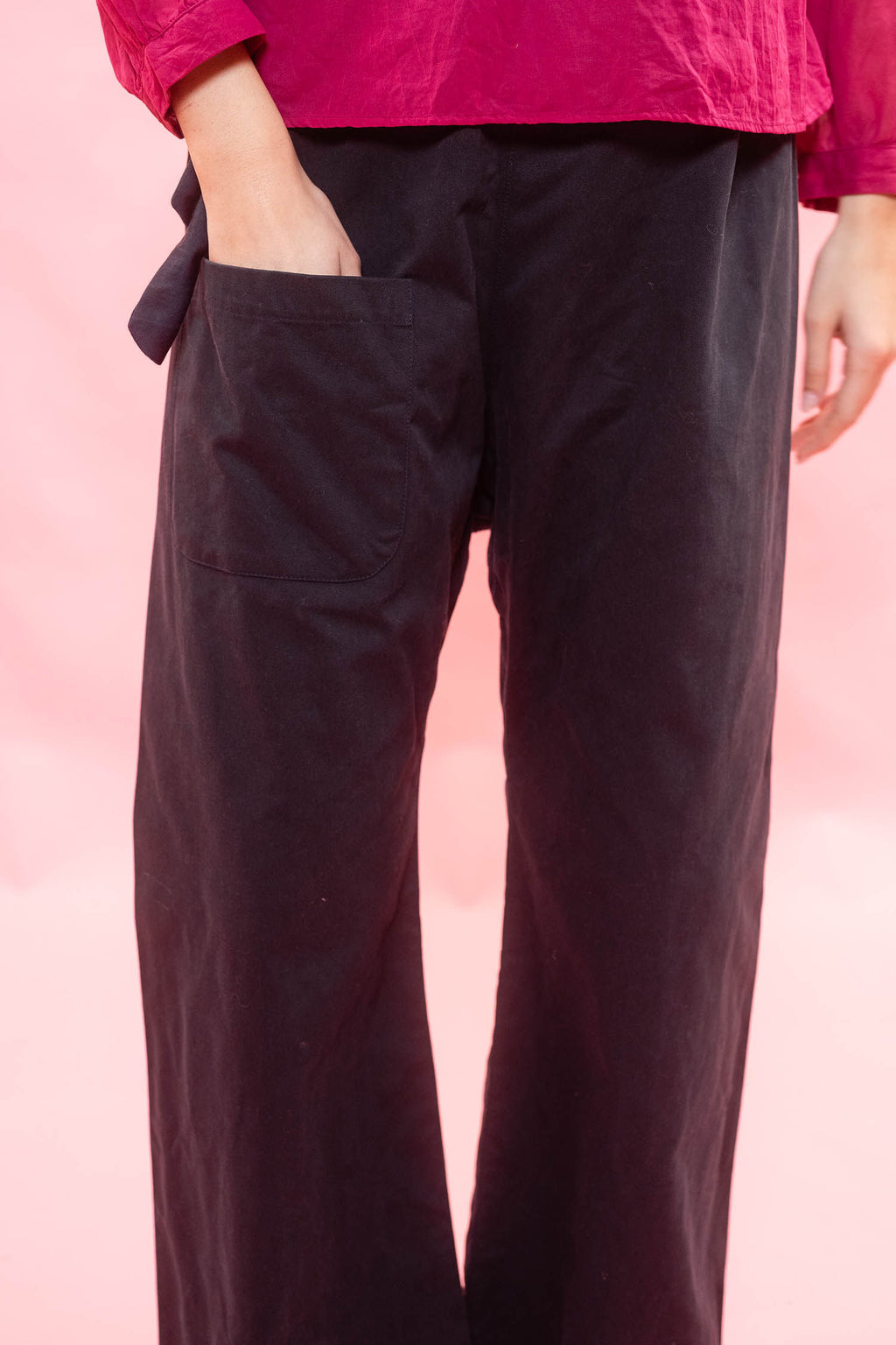A close-up of a person wearing dark navy wide-leg trousers with a side tie and a large patch pocket, paired with a bright pink top against a soft pink background.