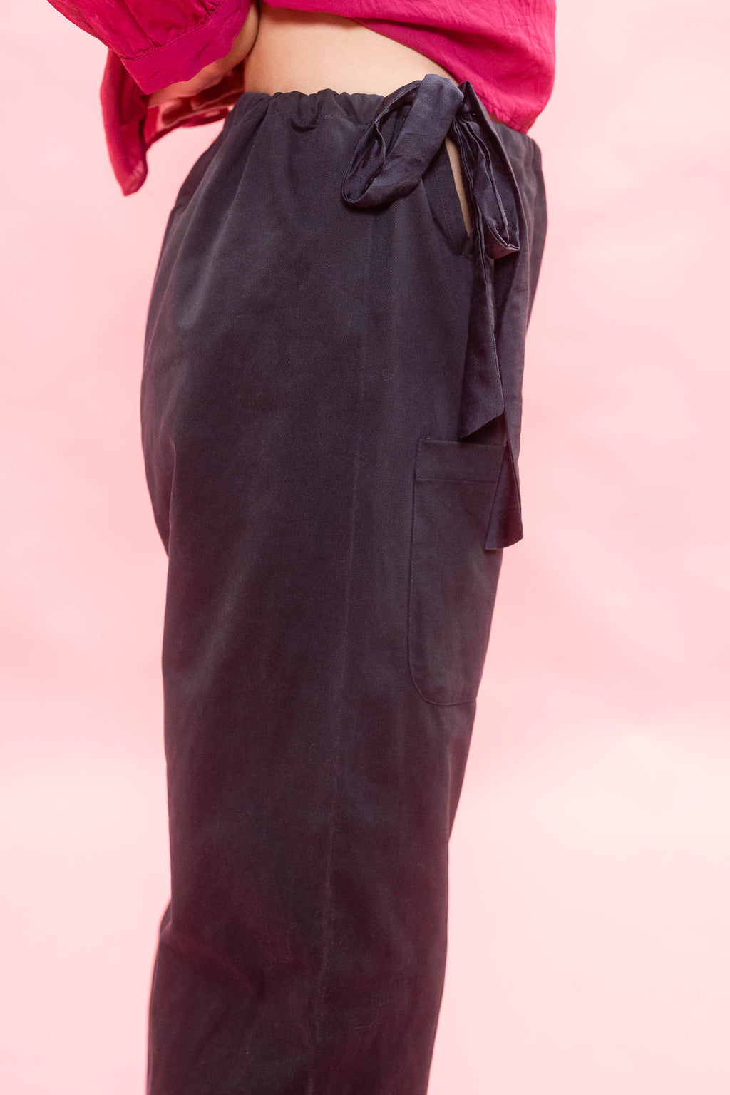 A close-up of a person wearing dark navy wide-leg pants with a side tie waist and a pink top, set against a solid pink background.
