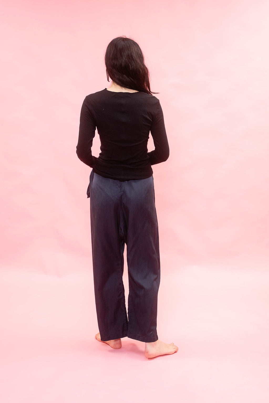 A person standing with their back to the camera, wearing a black long-sleeve top and dark navy wide-leg pants, set against a solid pink backdrop.