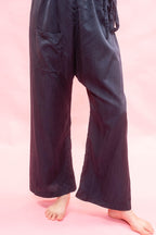 A person wearing loose, wide-leg pants in a very dark navy or near-black fabric, standing barefoot against a solid pink background. The pants have a drawstring waist and a side patch pocket.