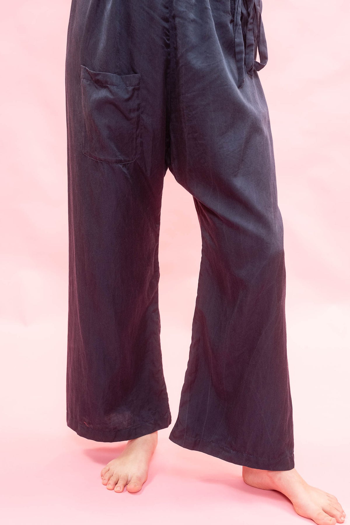 A person wearing loose, wide-leg pants in a very dark navy or near-black fabric, standing barefoot against a solid pink background. The pants have a drawstring waist and a side patch pocket.