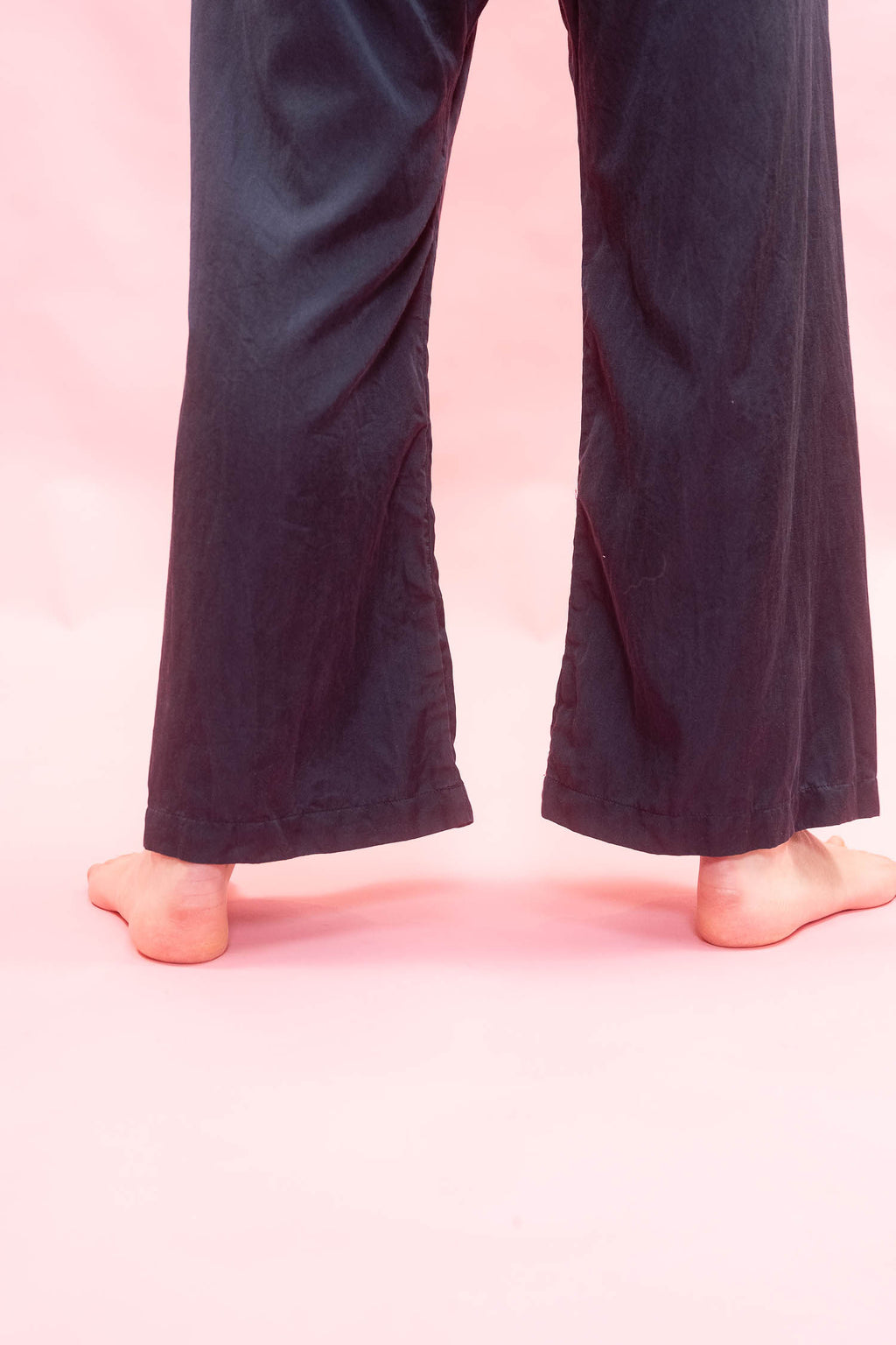A person's bare feet and lower legs wearing dark navy wide-leg pants against a solid pink background.